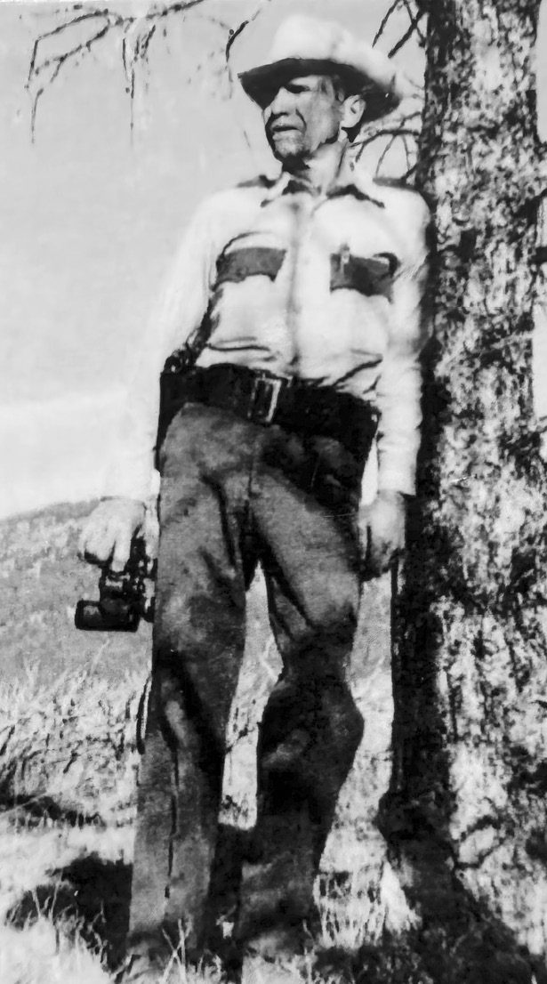 Wayne Fitzwater in warden clothes, binoculars in hand, leaning on a tree